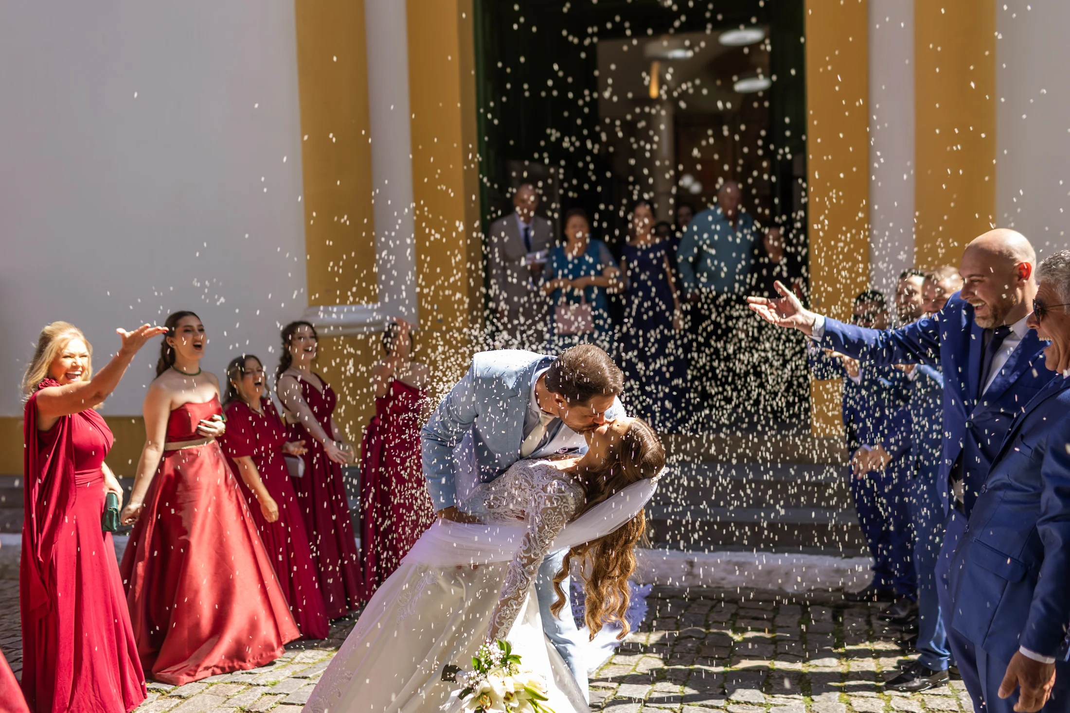Noivos se beijando sob uma chuva de arroz na saída da igreja, cercados pelos convidados